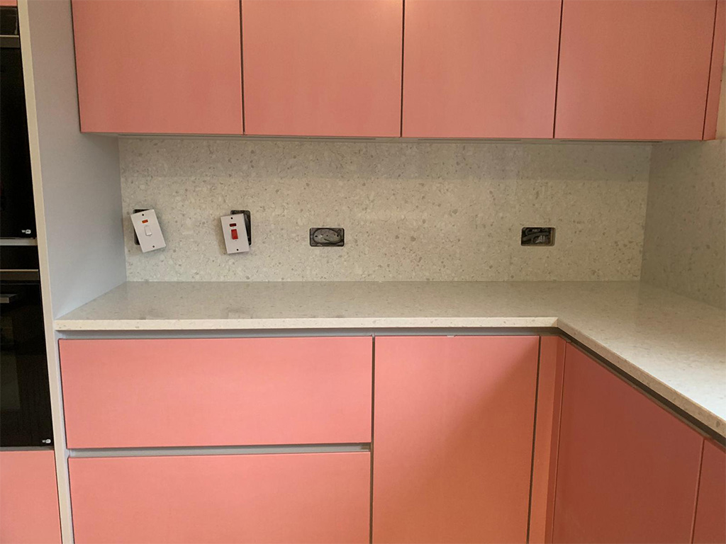 Speckled white quartz splashback with socket cut-outs, the sockets not yet refitted, between pink wall and base units
