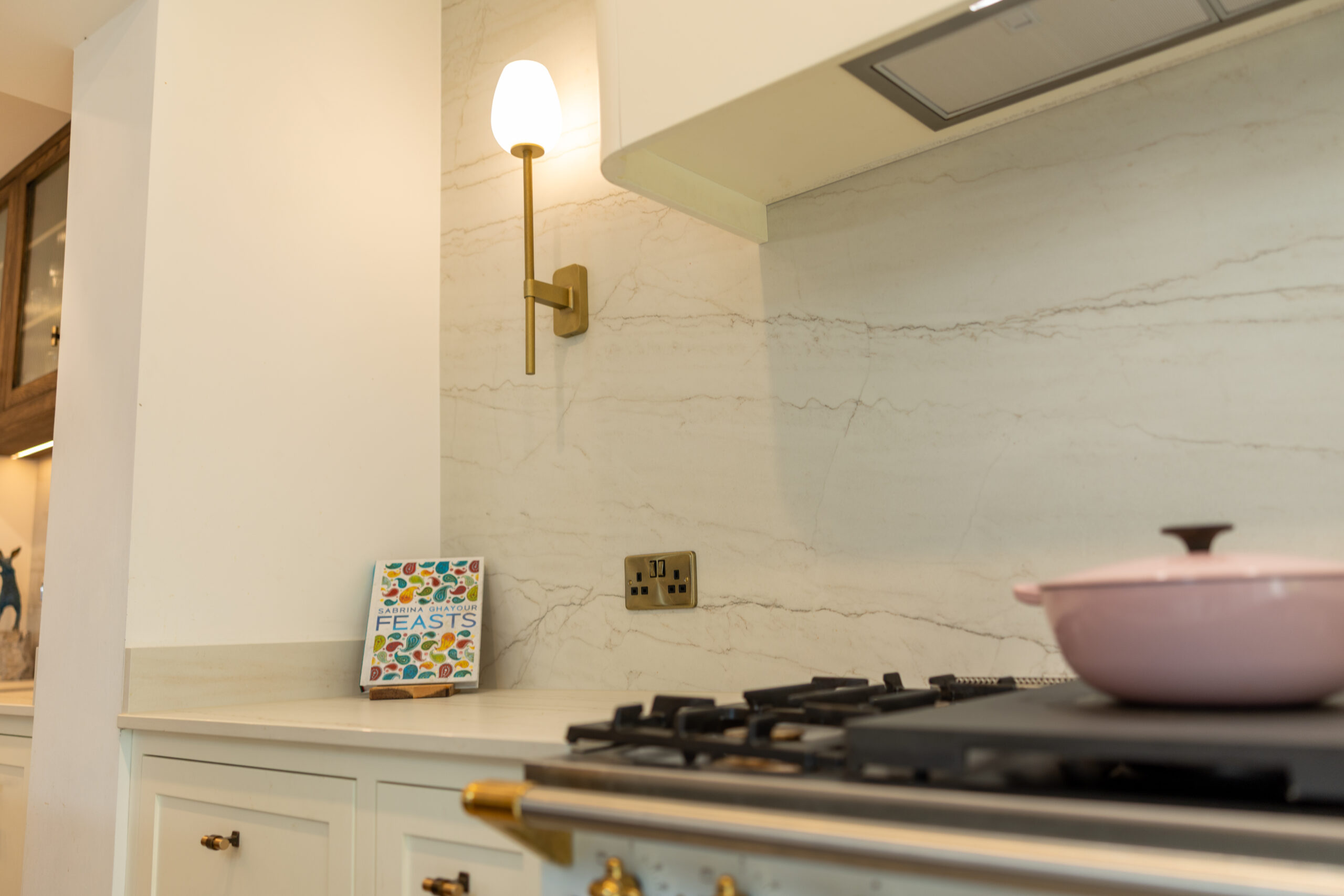 Full-height veined stone splashback behind a range cooker, rising to the extractor, with a brass socket set into the stone and a low upstand on the painted wall beside it